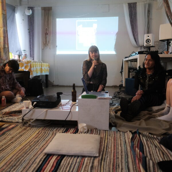 group of people in the interior of a studio, with a big screen in the background., One of them holding a mic. The vibe is chill and informal.